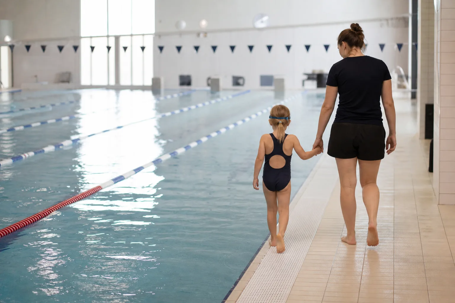 Barn i baddräkt går med vuxen längs kanten av simskolebassäng