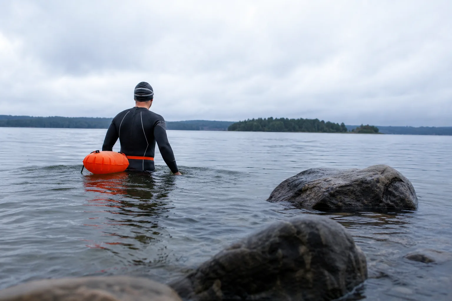 Simmare i våtdräkt vadar ut i sjö med orange simboj fäst runt midjan